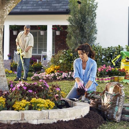A woman planting some bulbs in top soil, while a man uses a strimmer in the background.
