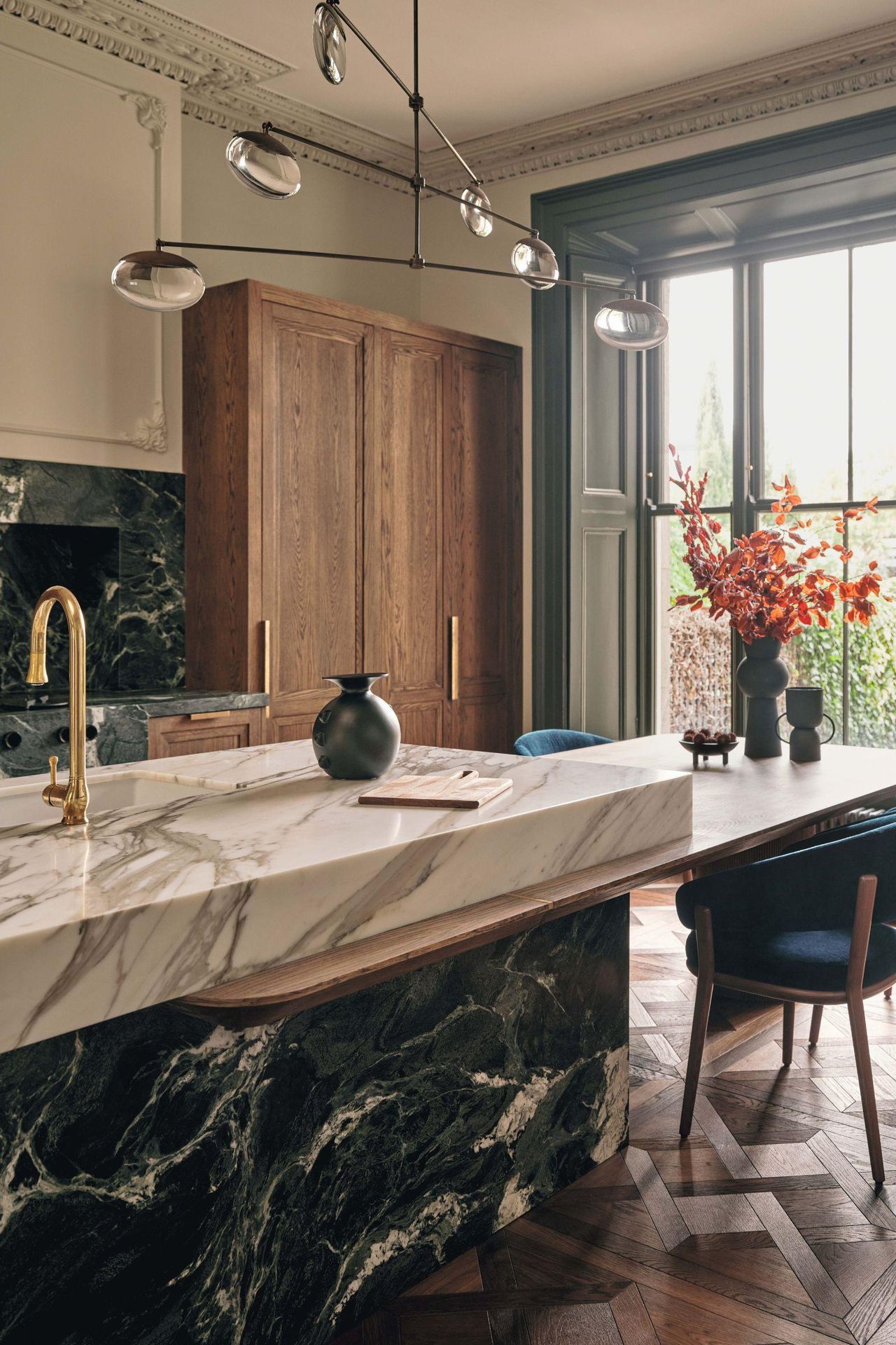 Left image: a wooden kitchen with dramatic dark marble island with a white marble countertop. There are large windows with dark trim and a sculptural light fixture hangs above the island. Right image: Elegant dining room featuring a large dark wood oval table, plush upholstered chairs, and a statement crystal chandelier. A mirrored cabinet with brass base stands against the wall, and tall windows are framed by soft, neutral curtains.