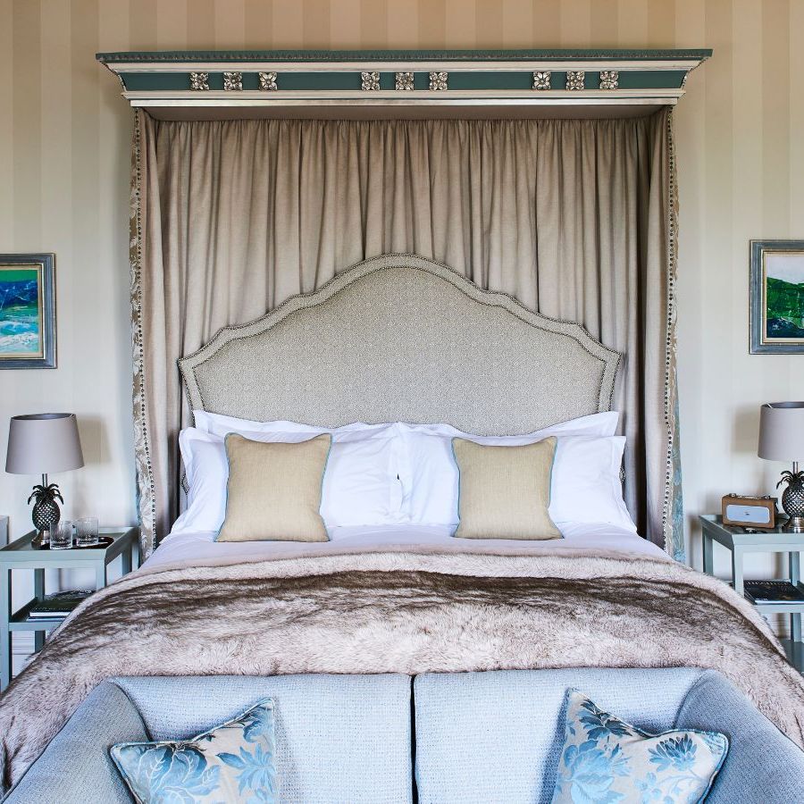 Bedroom with large double canopy bed and cream colored striped wallpaper. At the end of the bed is a pale blue sofa with matching pillows, and to either side of the bed are pale blue bedside tables with matching pineapple lamps.