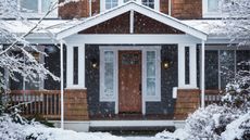 Exterior shot of a large home in the winter, with snow on peaked porch, front yard, path and falling. The front door is wooden as are the porch guard rails