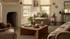 A cozy but bright cream living room with cream upholstered chairs, cuddle seats and mid-centrury wooden coffee table, side table and sideboard. There is a stone mantel and sheer window treatments in view, as well as a checkered area rug