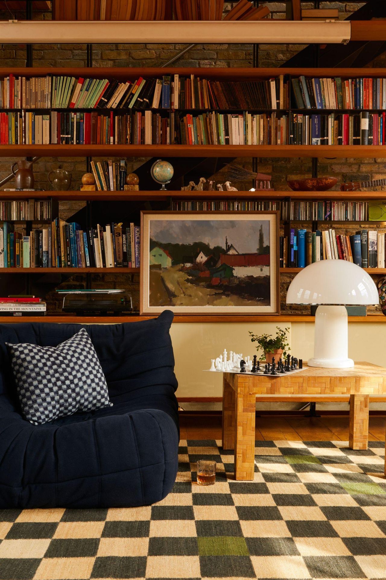 A cozy, mid-century modern living room featuring floor-to-ceiling brick bookshelves, a navy blue Ligne Roset Togo sofa, and a black-and-white checkered rug