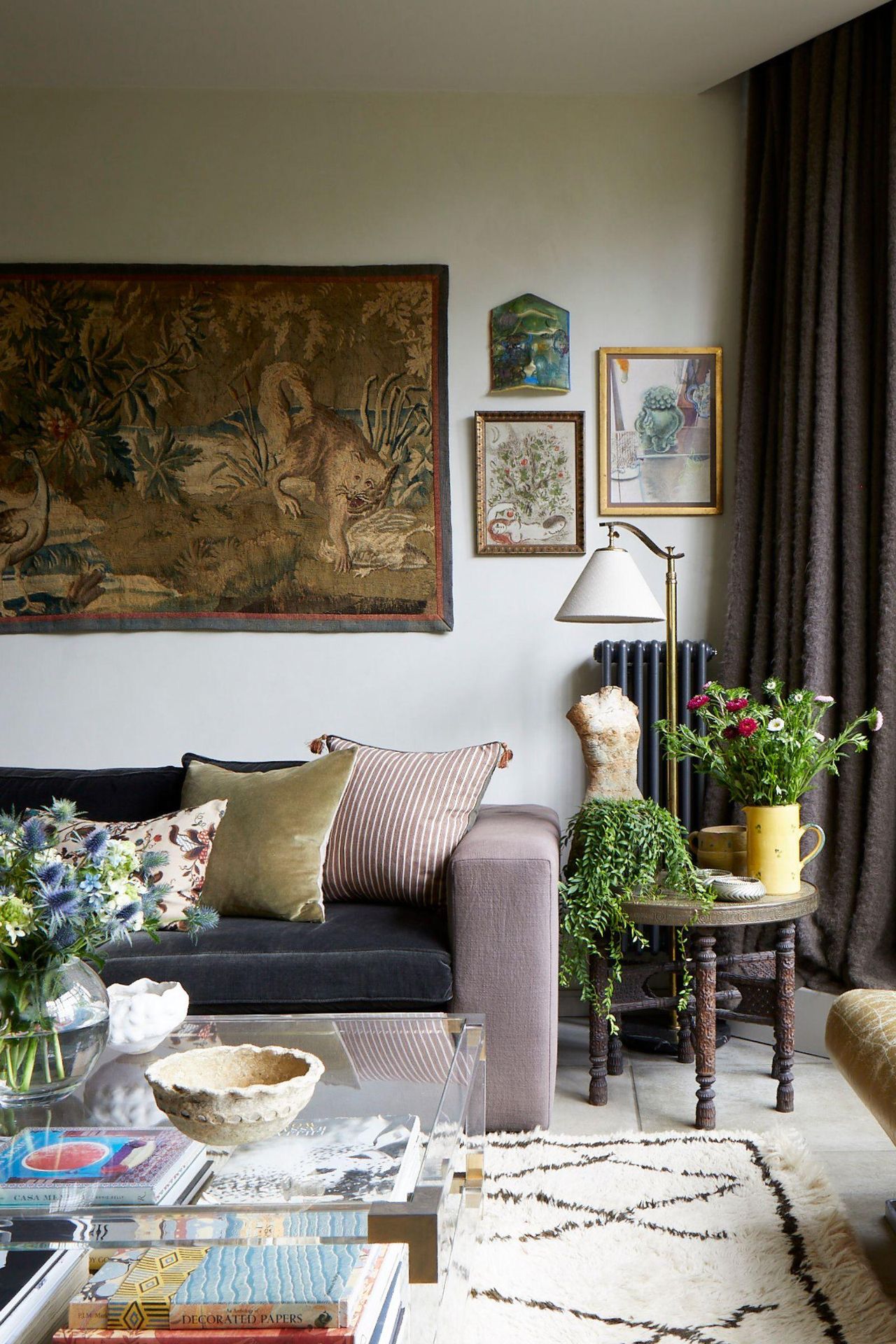 An eclectic, cozy living room featuring a velvet sofa with striped pillows, a large animal tapestry on the wall, and a glass coffee table layered with books and flowers