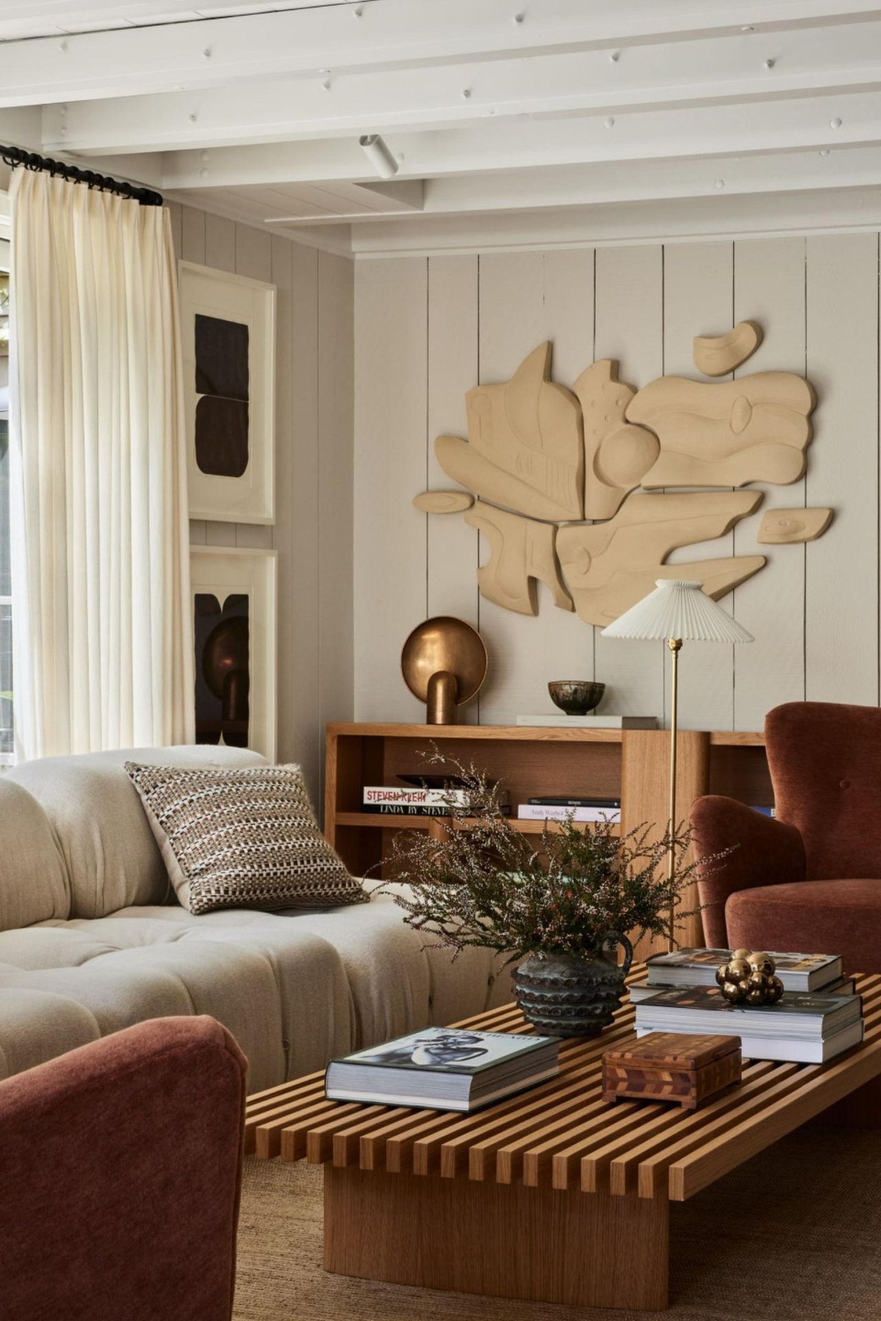 A bright, contemporary living space with cream wood-paneled walls, a slatted oak coffee table, and a sculptural tan wall installation above a terracotta armchair