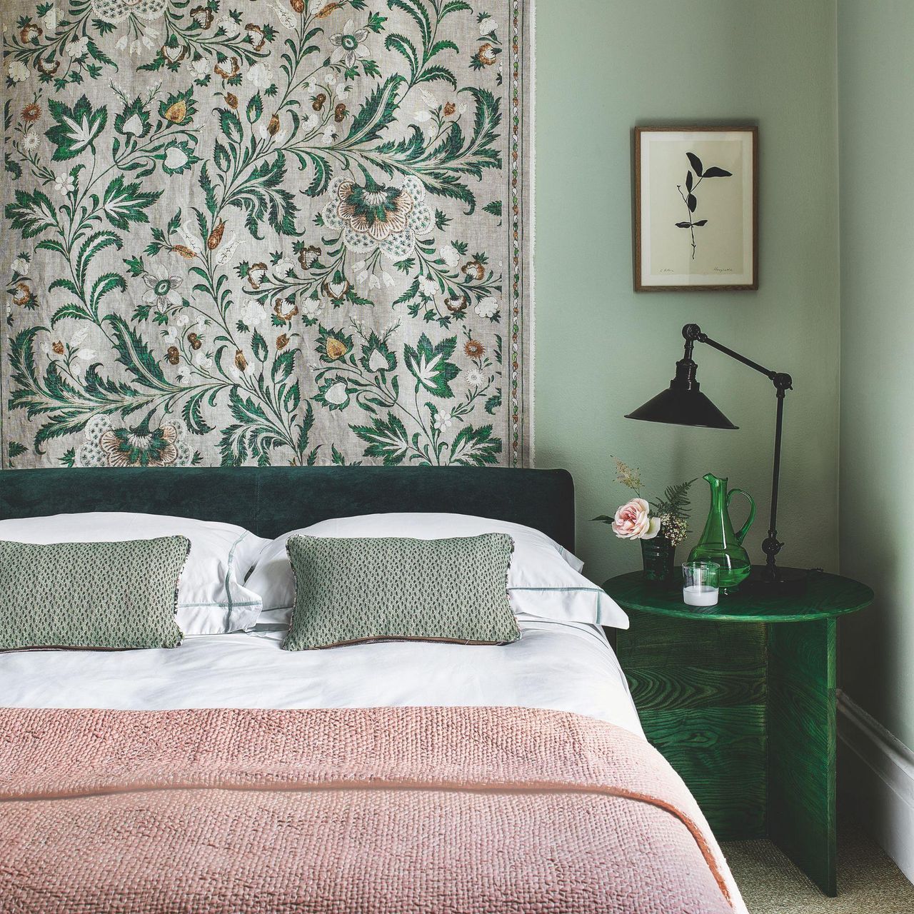 A green bedroom with a green velvet bed with white bedding, a pink blanket and green cushions, a patterned hanging tapestry behind the bed, a green bedside table with a green glass jug, pink flowers and a black lamp, with a small botanical artwork above. The walls are color drenched sage green.