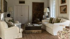 a traditional white living room with a large antiqu tapestry and vintage art, wall moldings, two white armchairs, a linen couch, green velvet fringed ottoman