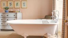 A pink wallpapered bathroom with a rolltop free standing tub. A distressed dresser in the background with eight gold framed pictures on the wall above it. 