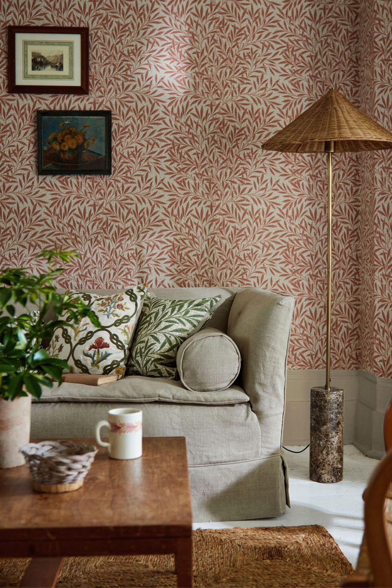 Two interiors showcasing traditional British style with patterned wallpaper by Morris &amp;amp; Co. The left image features a bedroom with dark green floral wallpaper leading into a coordinating bathroom, a striped headboard, and a vintage wooden side table. The right image shows a cozy living room with soft red leaf-patterned wallpaper, a neutral sofa with botanical cushions, a wooden coffee table, and a woven floor lamp, creating a warm and timeless look