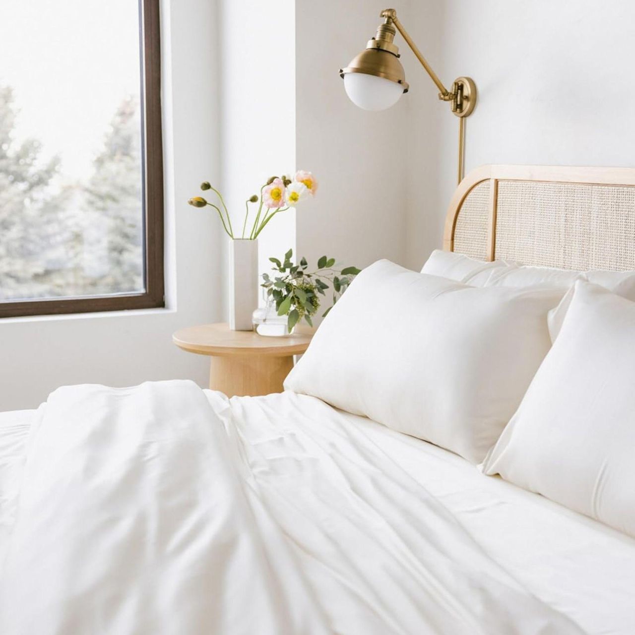 White Cozy Earth bedding on a rattan bed frame. Next to the bed is a circular wooden table with two white vases of flowers, a gold wall-mounted lamp, and a large window. The walls are white.