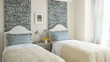 A white panelled twin bedroom with two twin beds with white headboards, with matching cream blankets, scalloped patterned blue pillows, and patterned wall tapestries behind each bed. Above the panelling is a strip of dark green wall, with white cornicing above.