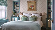 A bed with light blue linens and green decorative pillows set against a delicate floral trellis wallpaper. A teal built-in bookshelf sits to the right, filled with blue and white ceramics.