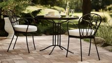 Whimsical wrought iron bistro table set topped with white wine and stemware, set atop of a brick patio overlooking a lush backyard