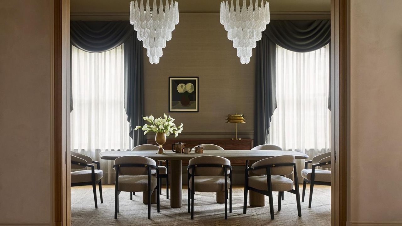 A symmetrical, sophisticated dining room viewed through a doorway. The space features a long wooden table surrounded by curved grey armchairs, two large tiered glass chandeliers, and tall windows with dramatic charcoal grey swags and sheer curtains