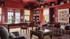 a vast home library clad in wood and stained red with a large antique desk, upholstered patterned couch, a tapestry on the wall and layered lighting
