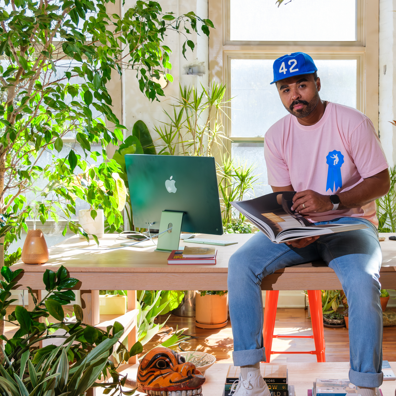 Man in jeans and pink t-shirt sat on office desk surrounded by large, topical indoor plants